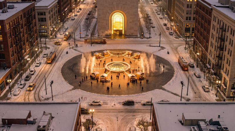 The Square, after dark: The same square at night: the warm circle lit by the glowing oculus and strings of lanterns, fountains steaming, people out in the cold, snow glowing on every roof.