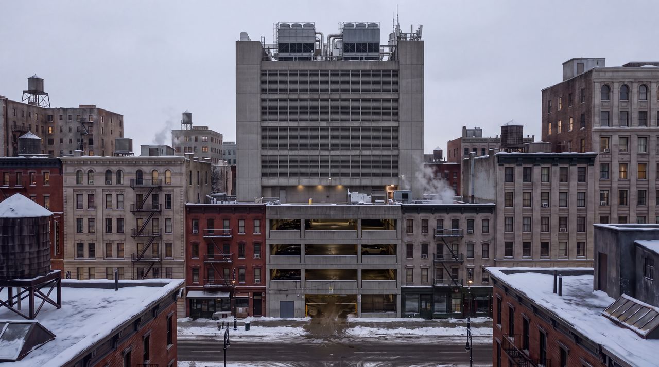 Today: The same view as it is now: a squat windowless gray concrete tower with louvered mechanical floors and a roof crowded with cooling units, a parking garage at its base, under an overcast sky.