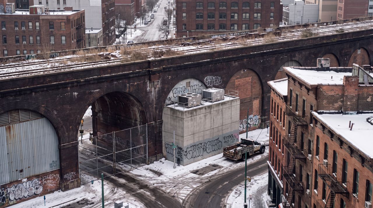 Today: The same street as it is now: a derelict brick viaduct with weeds on top, its arches bricked up or fenced, graffiti, a gray windowless box beside it, dirty snow, under an overcast sky.